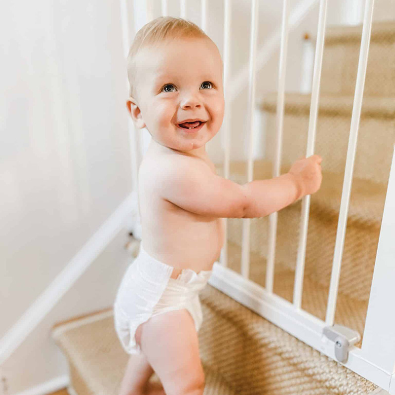 A smiling baby standing near a safety gate on a staircase, indoor home setting, Ocean City Moms.