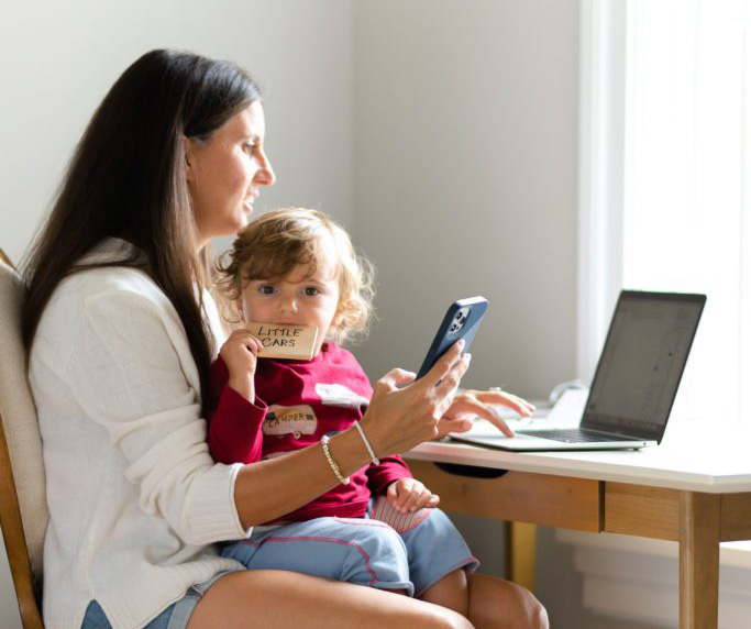 A woman and young girl using a smartphone and laptop at home, bright natural light.